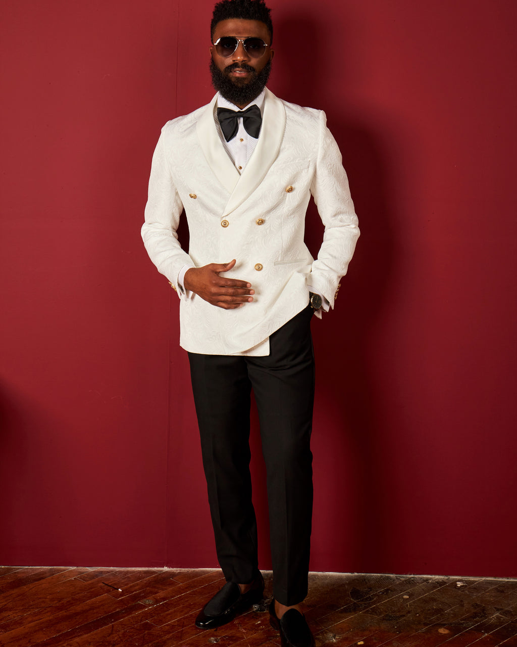 Man in a white double-breasted suit with black pants and bow tie against a red wall.