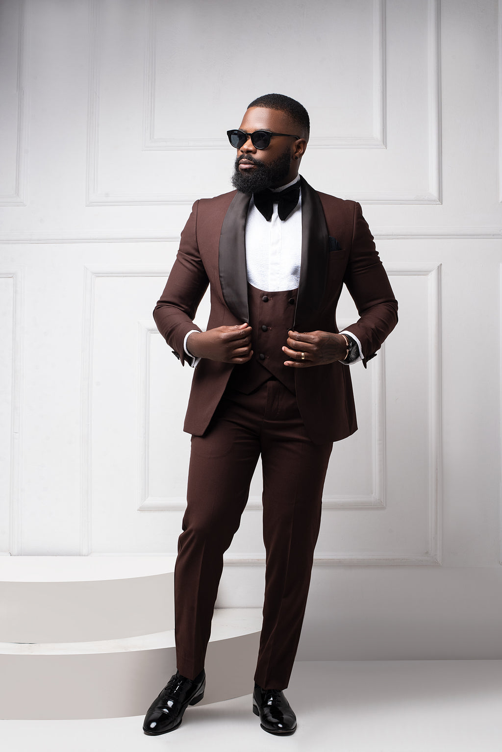 Man in a brown tuxedo with a white shirt and black bow tie against a white paneled wall.