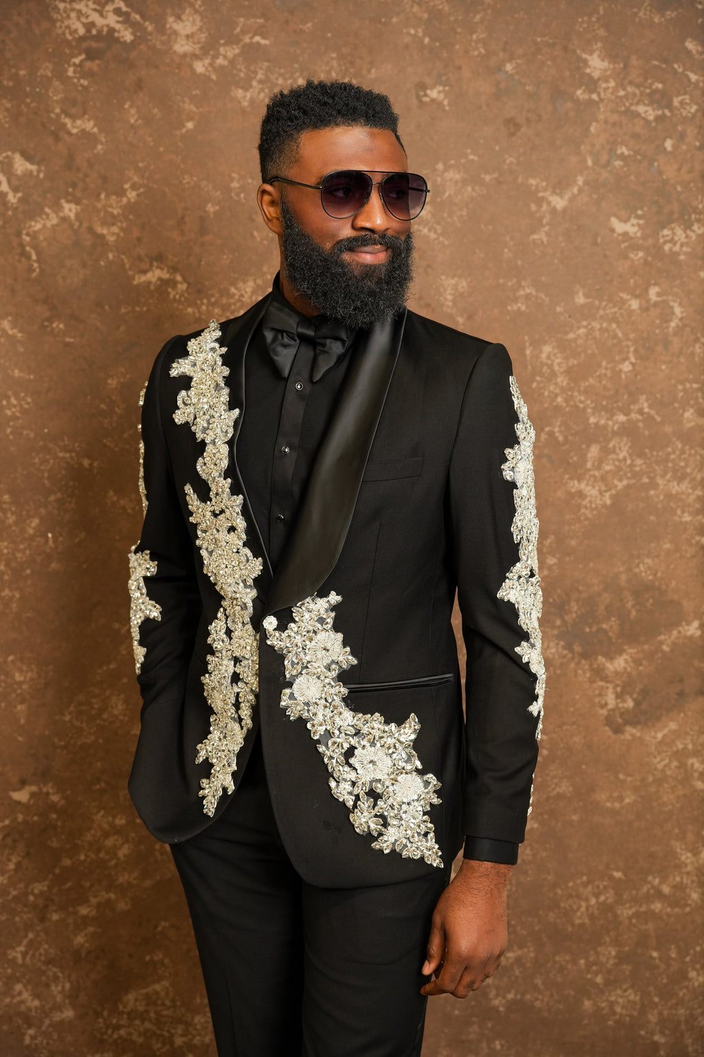 Man wearing a black suit with white floral embroidery against a brown textured background