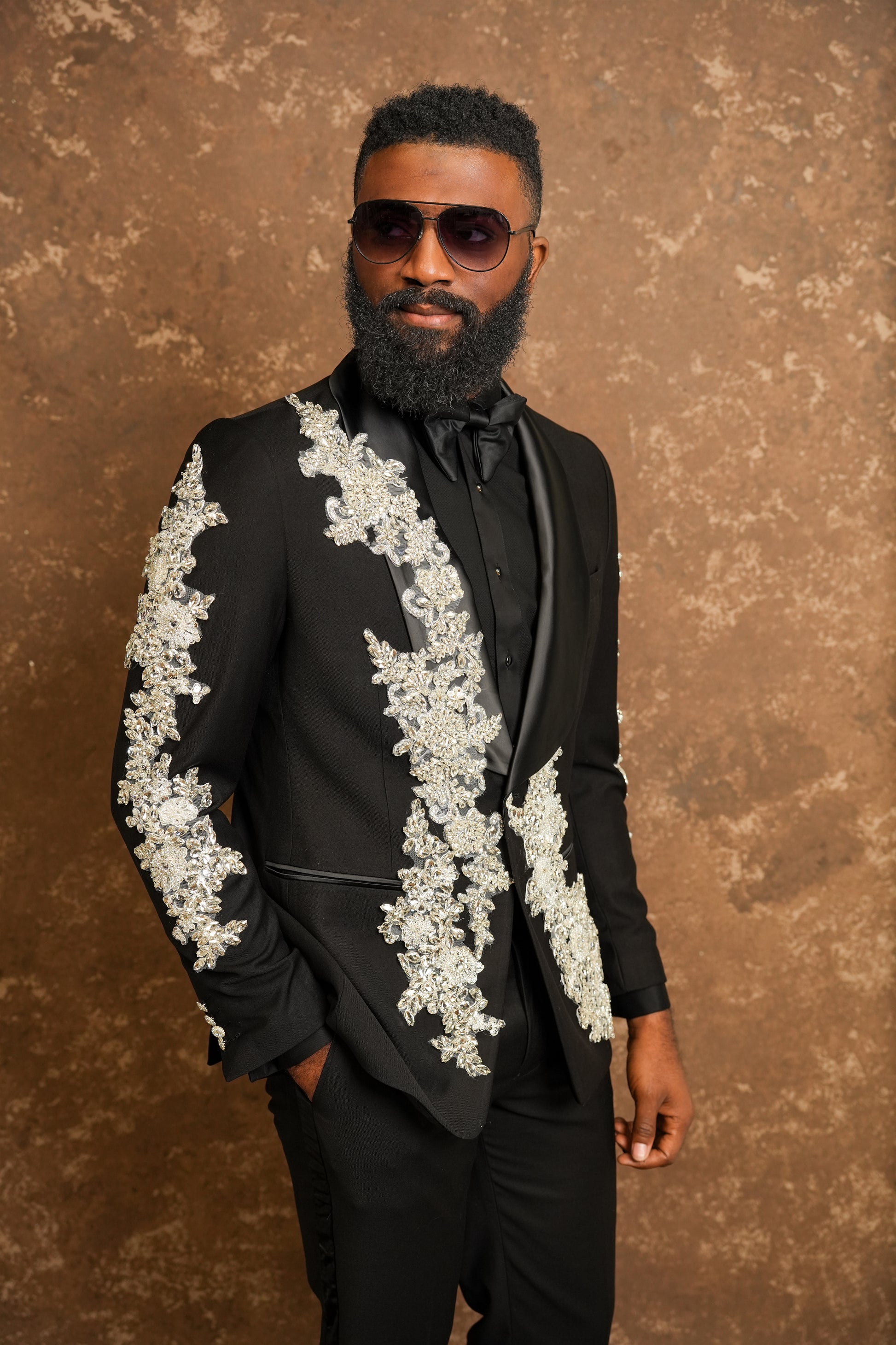 Man wearing a black suit with white lace details against a brown textured background