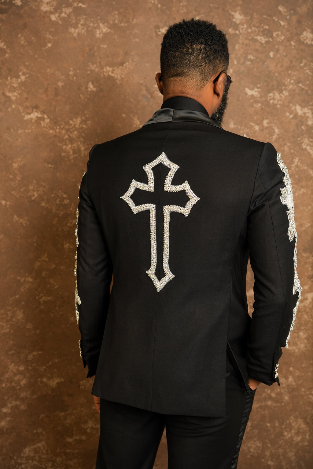 Man wearing a black jacket with a white cross design on the back against a brown textured wall.