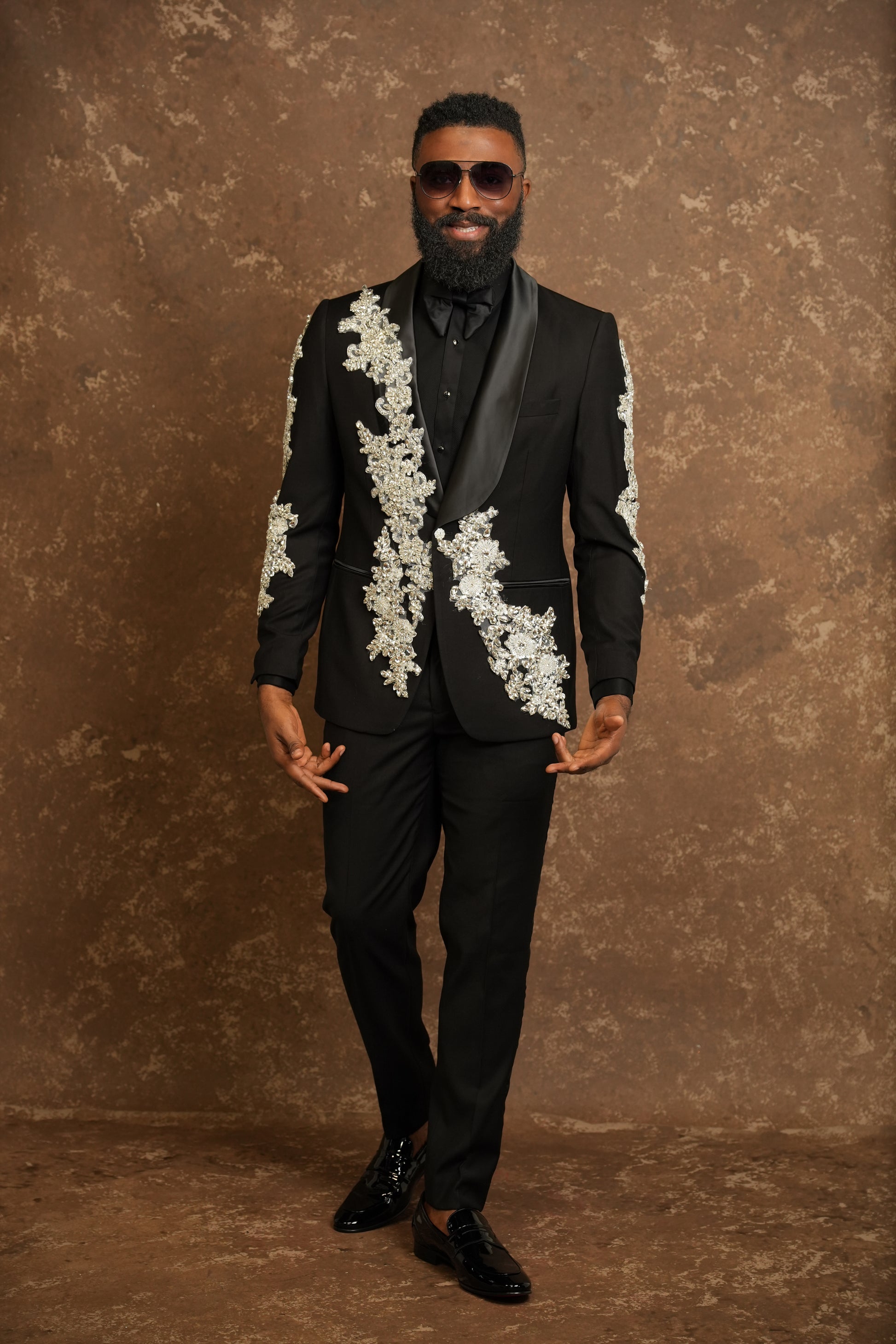 Man wearing a black suit with white floral embroidery against a brown textured background