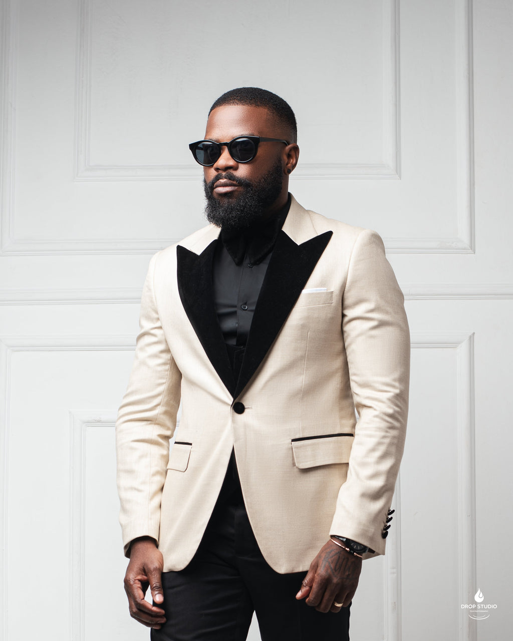 A man with a full beard and sunglasses wearing a tuxedo jacket with wide black velvet lapels over a black shirt and black trousers.