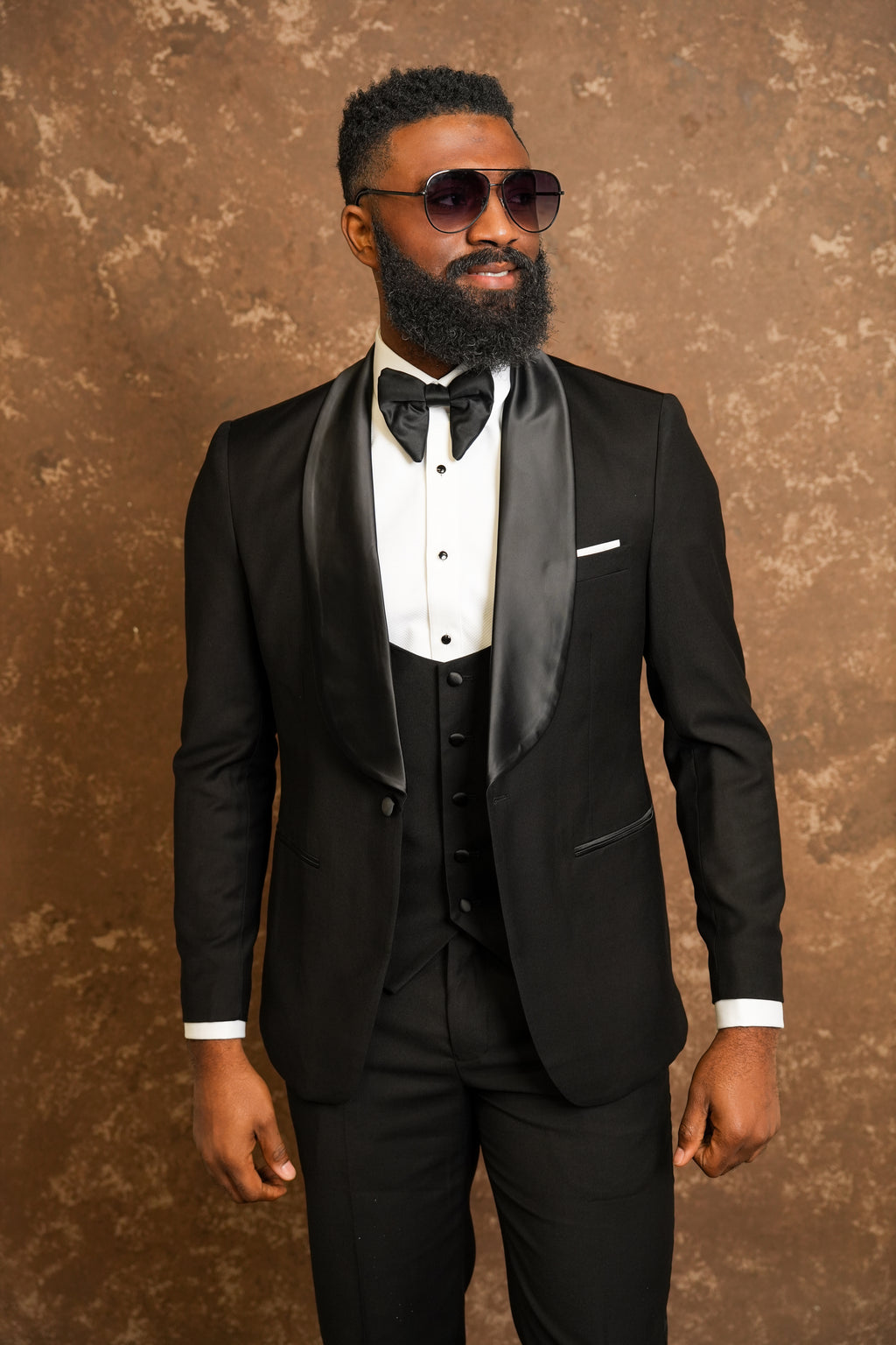 Man wearing a black tuxedo with a bow tie against a brown background