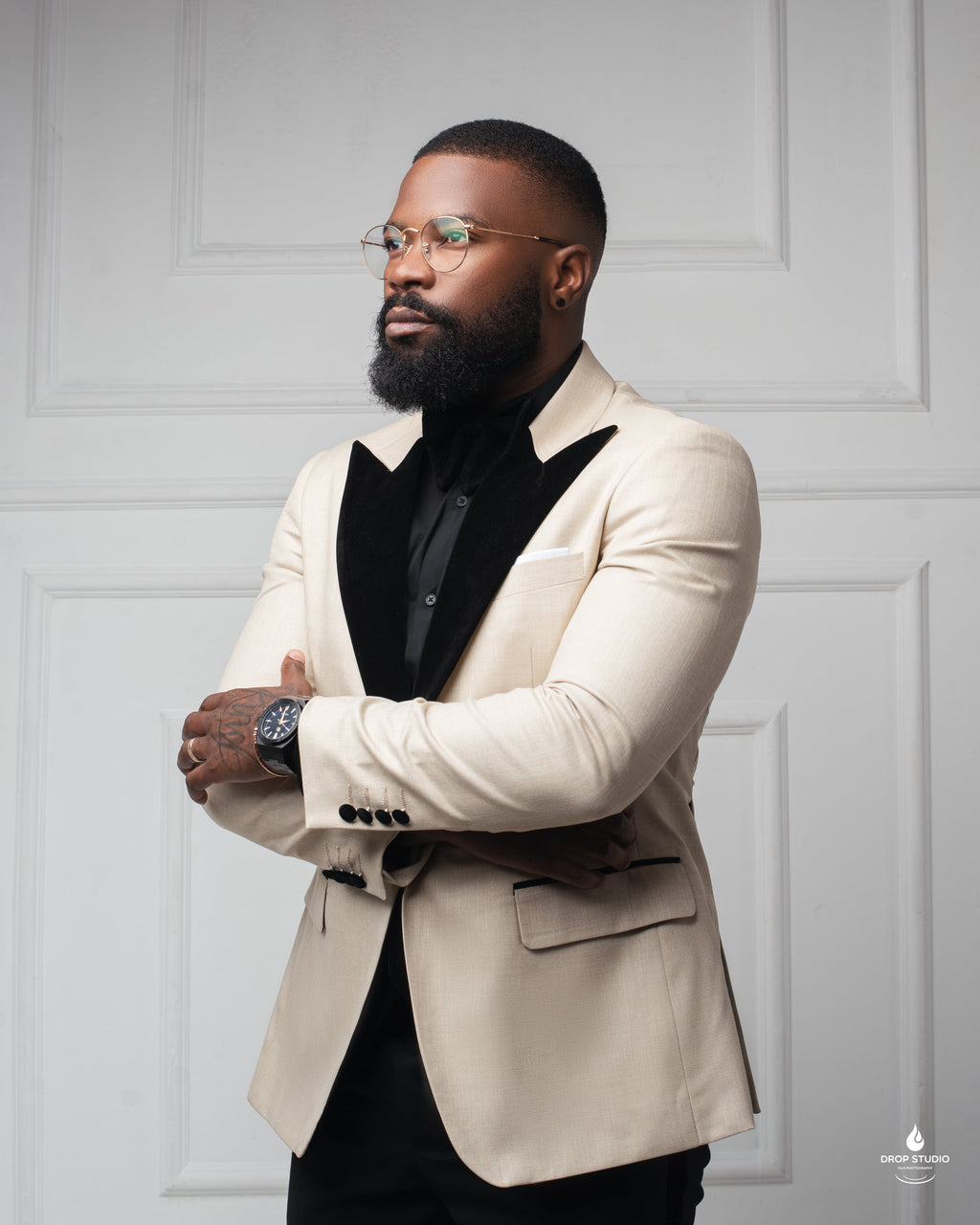 A bearded man wearing rimless glasses and a savannah black tuxedo over a black shirt, standing with arms crossed against a white paneled wall.