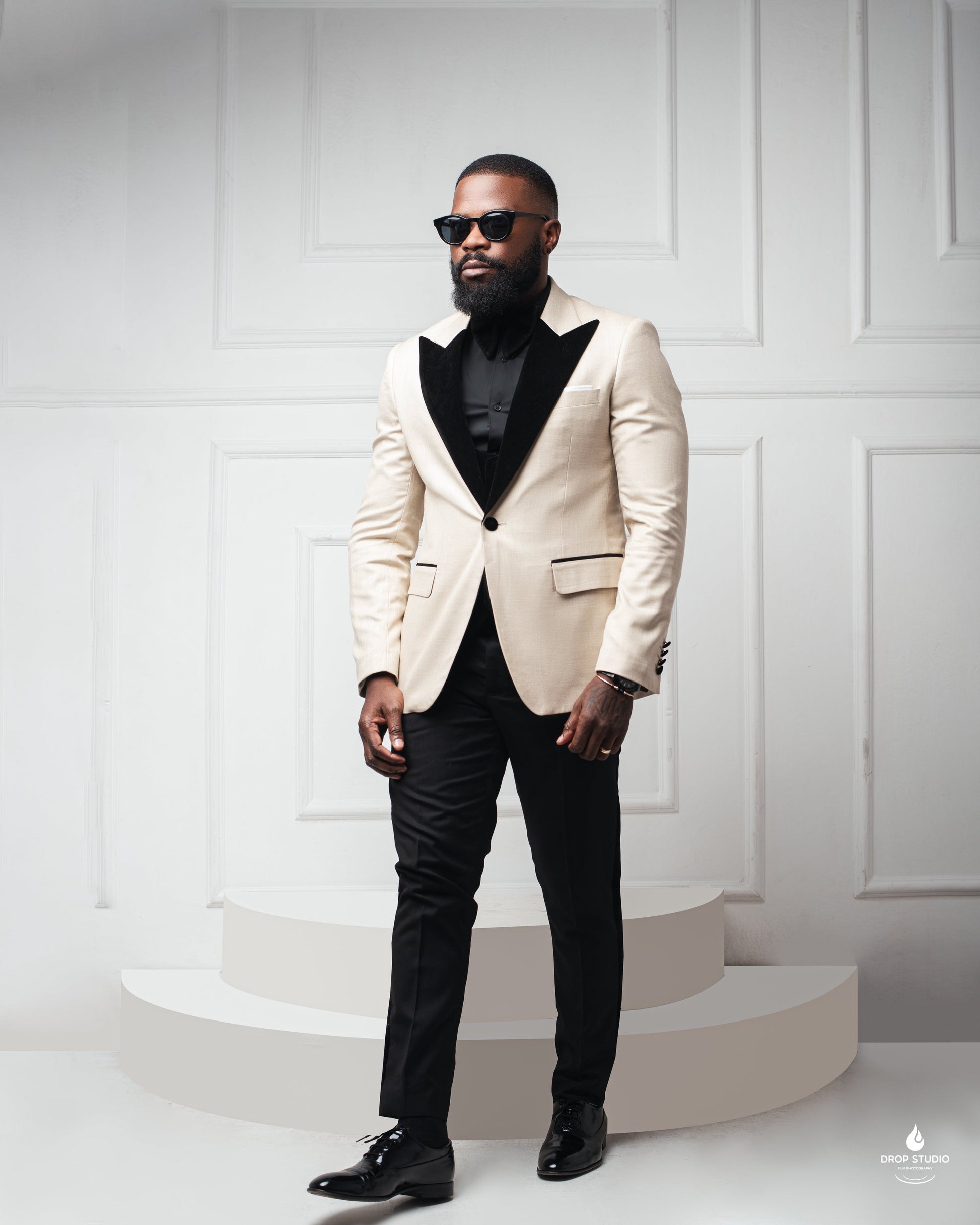 A sharply dresse  man with a full beard and sunglasses stands on a white stepped platform against a clean, paneled white background